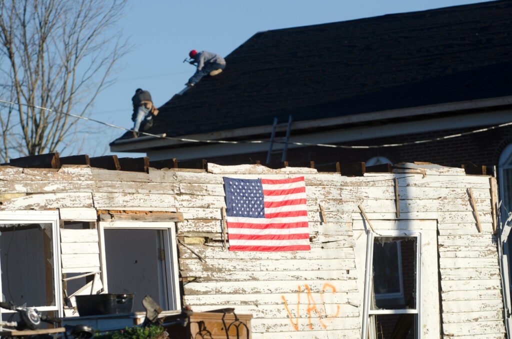 Workers fix the roof of a church while an American flag is hanging on the wreckage of a home damaged